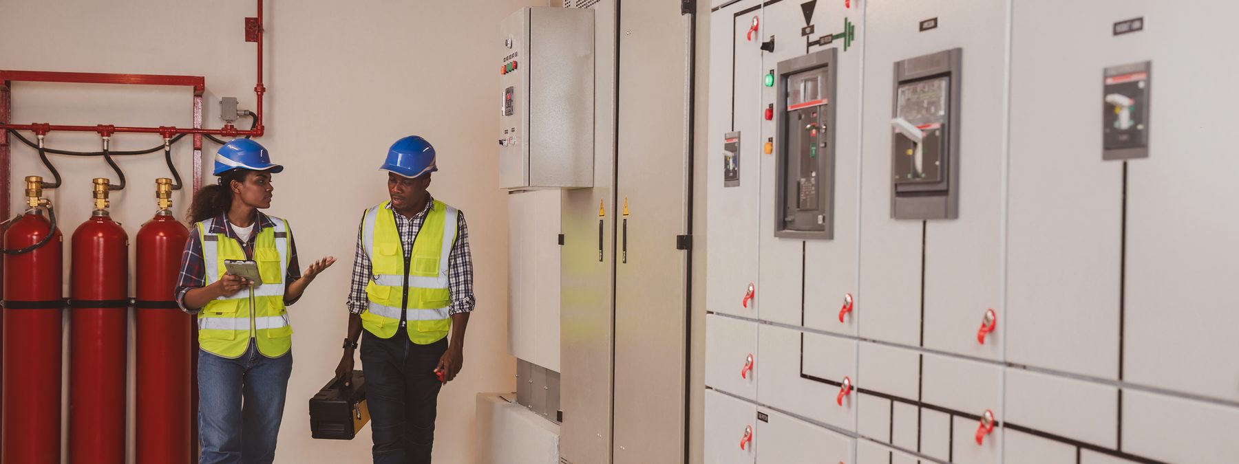 Two engineers in hard hats and high-visibility vests walking a switchgear room, reviewing equipment.