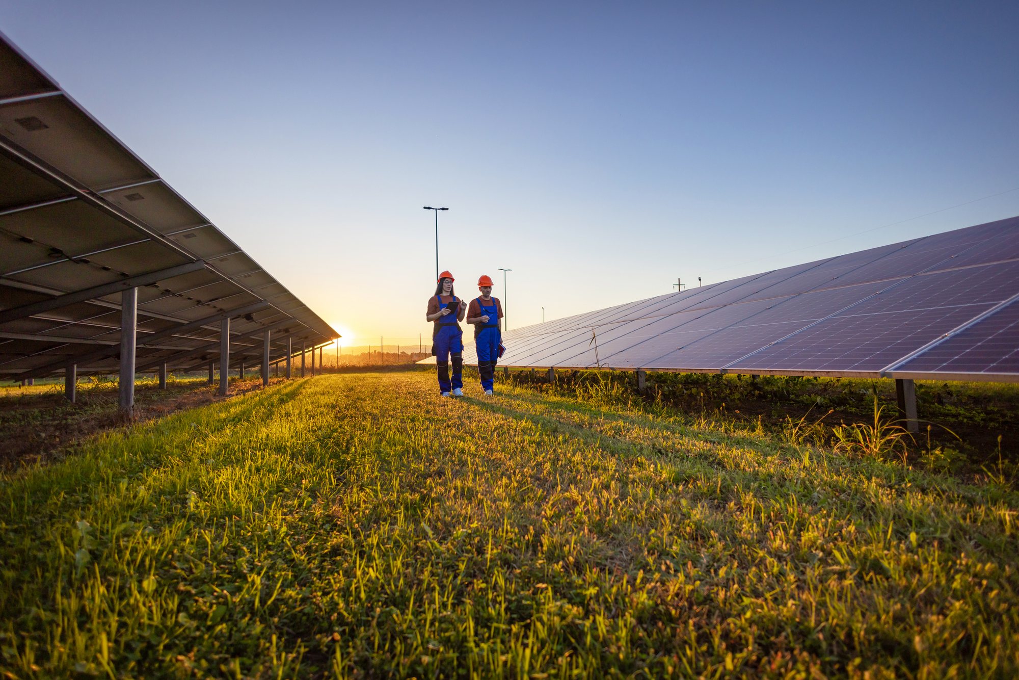 Two energy workers in hard hats walking a solar array at sunset.